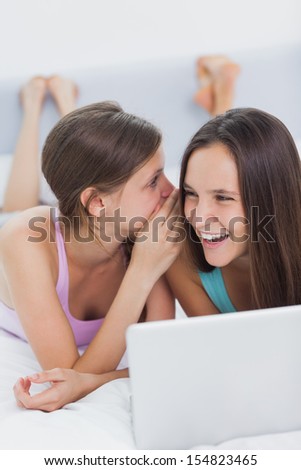 Happy girls lying in bed with laptop and telling secrets