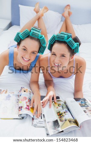 Happy friends in hair rollers lying in bed looking and smiling at camera at sleepover
