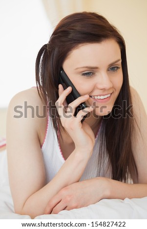 Happy brunette calling someone with a mobile phone lying on a bed in a bedroom