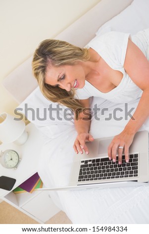 Cheerful mature blonde woman typing on a laptop and lying on her bed in a bedroom