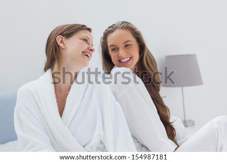 Happy friends wearing bathrobe sitting on bed and talking