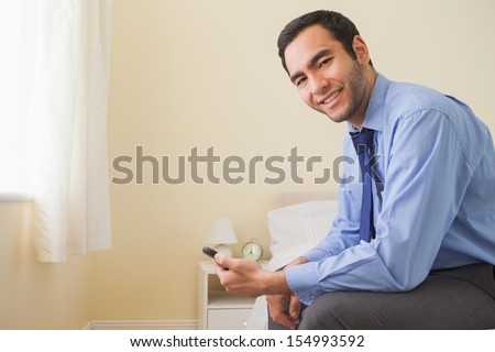 Pleased man looking at camera sitting on a bed using a mobile phone in a bedroom at home