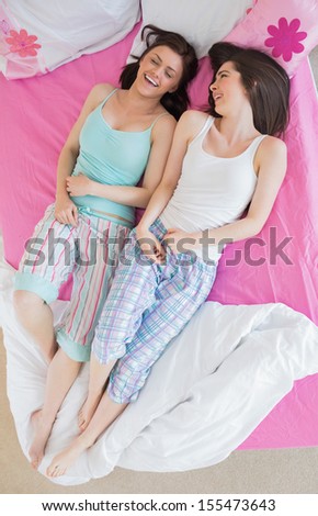 Friends in pajamas lying on bed and laughing in bedroom at home