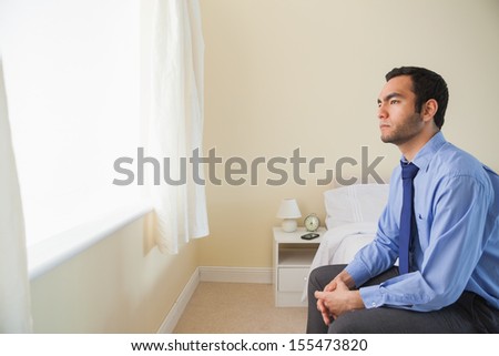 Unsmiling man looking out the window sitting on his bed in a bedroom at home