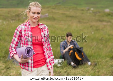 Happy woman carrying camping mat with boyfriend packing backpack behind her