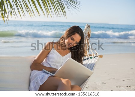 Pretty brunette sitting on hammock with laptop on the beach