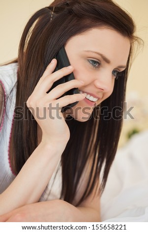 Smiling brunette calling someone with a mobile phone lying on a bed in a bedroom