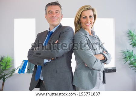 Two happy mature businesspeople looking at camera standing firmly back to back with crossed arms at office