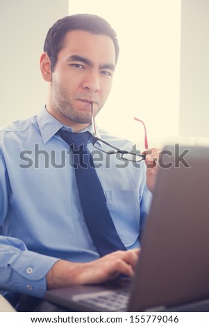 Irritated man biting his eyeglasses and using a laptop sitting on a sofa looking at camera