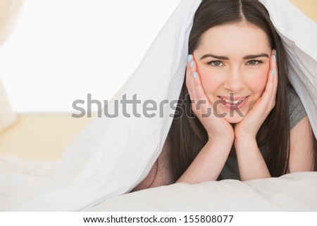 Happy brunette looking at camera lying hidden in her bed in a bedroom
