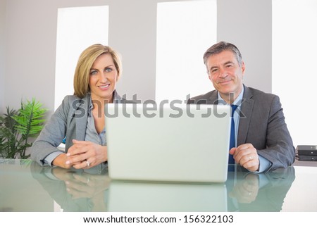 Two smiling mature business people looking at camera behind an opened laptop at office