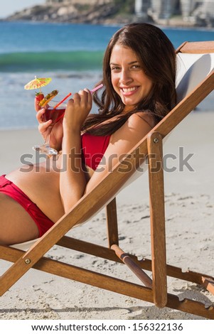 Cheerful pretty woman on the beach drinking cocktail while relaxing on her deck chair