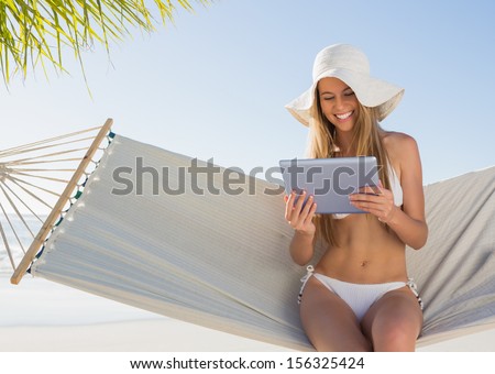 Cheerful blonde sitting on hammock using tablet pc at the beach