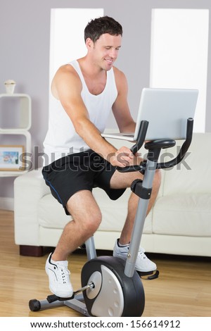 Happy sporty man exercising on bike and using laptop in bright living room