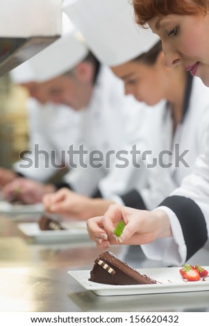 Team Of Young Chefs In A Row Garnishing Dessert Plates In Commercial Kitchen