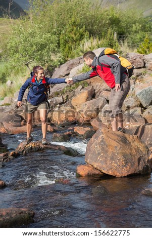 Man helping his girlfriend to cross a river carrying backpacks on a hike