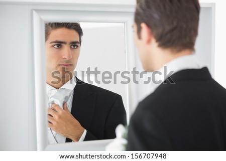 Serious handsome bridegroom looking in mirror straightening his tie