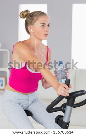 Sporty stern blonde training on exercise bike listening to music in bright living room