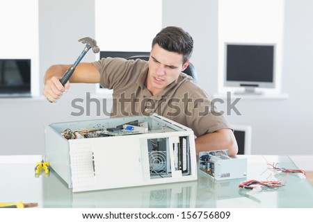 Attractive angry computer engineer destroying computer with hammer in bright office