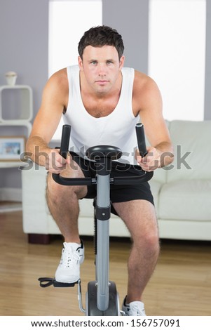 Handsome sporty man exercising on bike looking at camera in bright living room