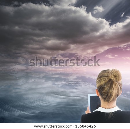 Composite image of blonde businesswoman holding new tablet in front of ocean and cloudy sky