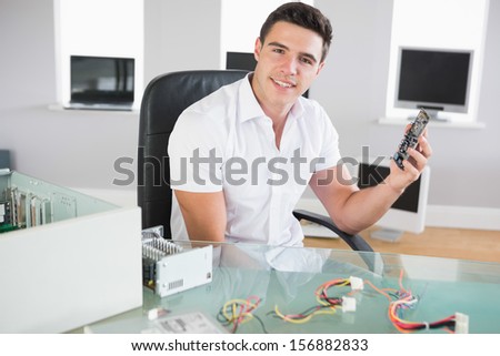 Attractive cheerful computer engineer sitting at desk holding hardware in bright office