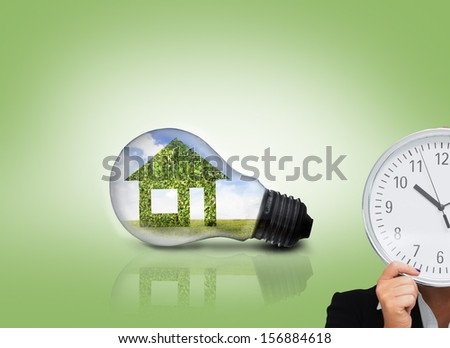 Composite image of businesswoman in suit holding a clock with huge light bulb showing green house in background