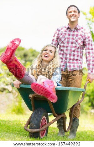 Smiling man pushing his laughing girlfriend in a wheelbarrow in sunny garden