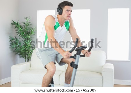 Stern handsome man training on exercise bike listening to music in bright living room