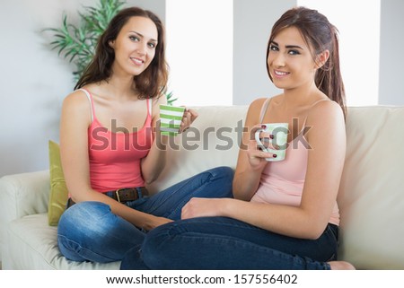 Two nice women sitting on a couch looking at the camera having coffee