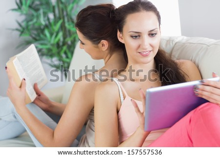 Two pretty women sitting on back to back on the couch with a book and a tablet