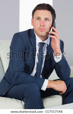 Unsmiling handsome businessman phoning and sitting on couch in bright living room