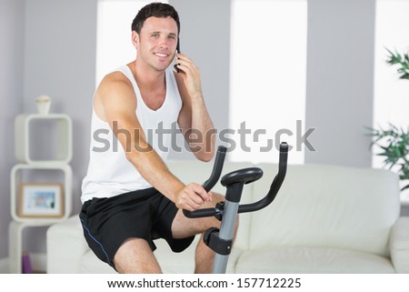 Cheerful sporty man exercising on bike and phoning in bright living room