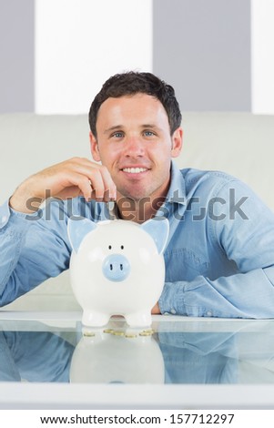 Smiling casual man putting coin in piggy bank in bright living room