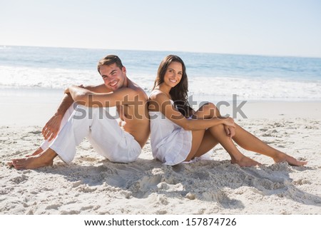 Couple sitting back to back on the sand smiling at camera at the beach