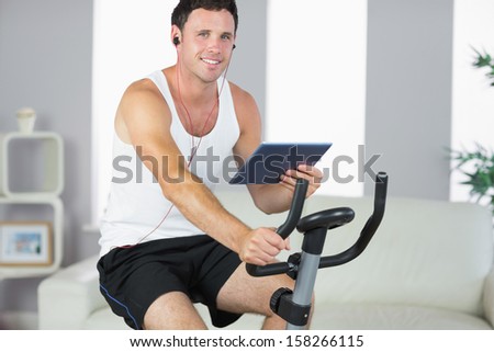 Smiling sporty man exercising on bike and holding tablet in bright living room