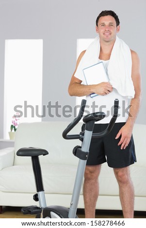 Cheerful sporty man standing behind exercise bike holding tablet in bright living room
