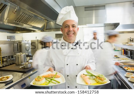 Mature head chef presenting proudly two dinner plates standing in a busy kitchen