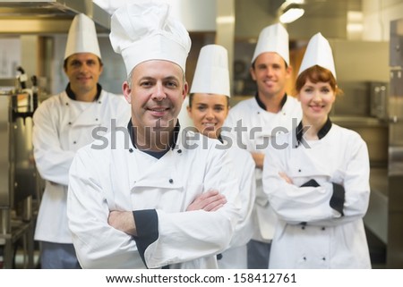 Five chefs wearing uniforms posing in a kitchen with crossed arms