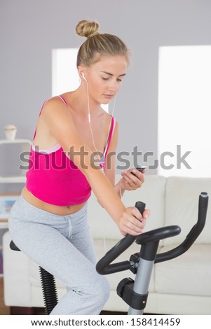 Sporty serious blonde training on exercise bike listening to music in bright living room