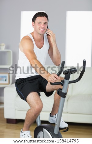 Content sporty man exercising on bike and phoning in bright living room