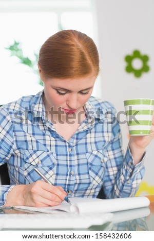 Redhead writing on notepad at her desk and holding coffee working from home