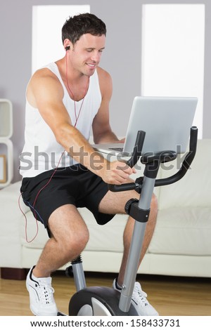 Sporty man with earphones exercising on bike looking at laptop in bright living room