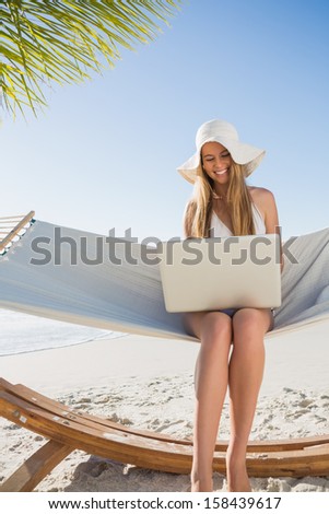 Cheerful blonde sitting on hammock using laptop at the beach