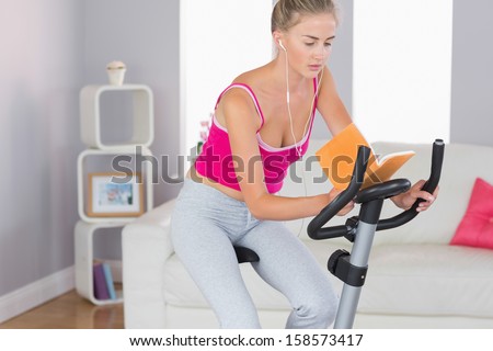 Sporty unsmiling blonde training on exercise bike reading a book in bright living room