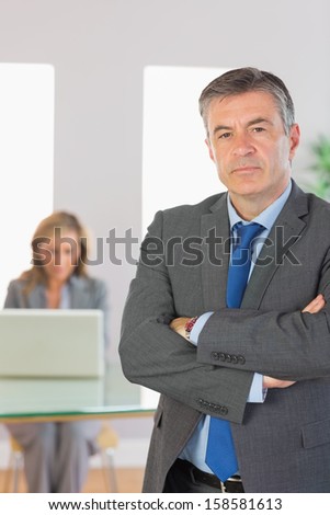 Irritated mature businessman looking at camera crossed arms with a blonde businesswoman working on laptop on background at office
