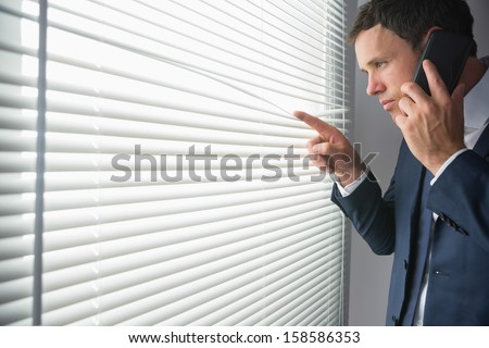 Unsmiling handsome businessman looking through roller blind phoning in dark room