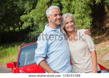 Happy mature couple posing by their red convertible on bright day