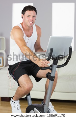 Sporty content man with earphones exercising on bike and holding laptop in bright living room