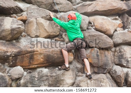 Determined man scaling a huge rock face wearing a red helmet and green jumper